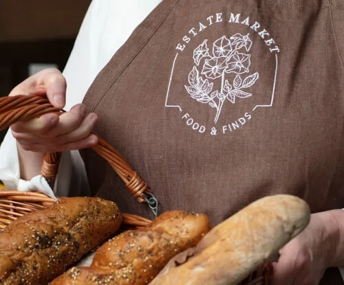 A person in a brown apron holding a basket of assorted breads.